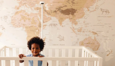 A toddler with curly hair leans against a white crib, set against a world map wallpaper featuring geographical details with nanit pro baby monitor with floor stand in background