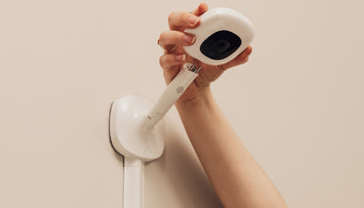 A close-up shot of a person's hand holding Nanit Baby Monitor with a black lens. Wall Mount is attached to a white, adjustable arm and base, against a plain light-colored wall.