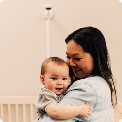 mom holding baby with nanit pro camera + wall mount and crib on the background