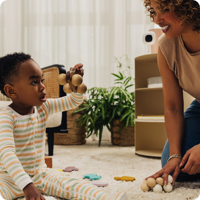 mom looking at toddler while toddler is holding a toy and nanit pro camera + flex stand is on bookshelf looking at mom and toddler